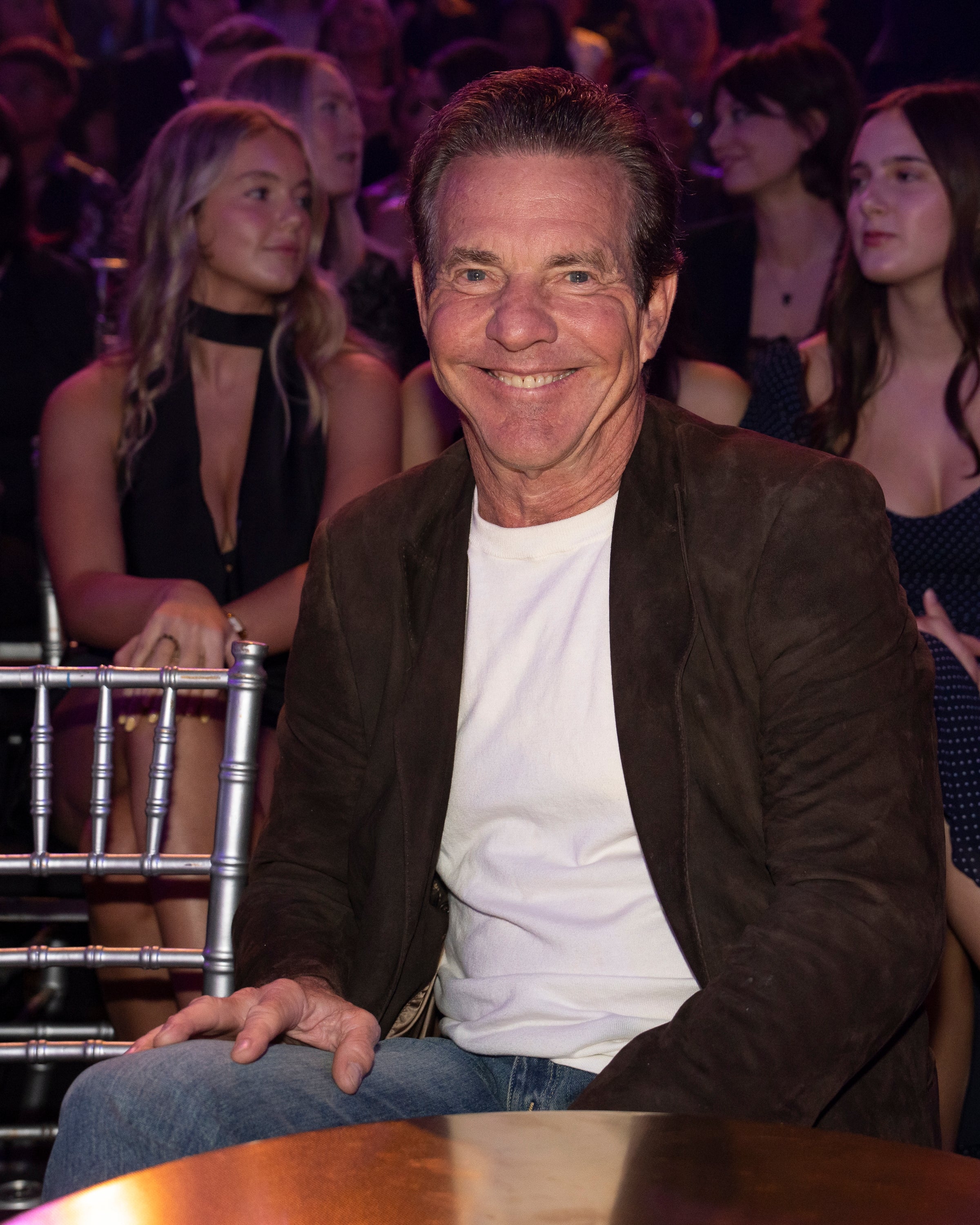 A smiling antheral successful a casual achromatic garment and acheronian blazer sits connected a seat astatine a nationalist event, with an assemblage successful the background