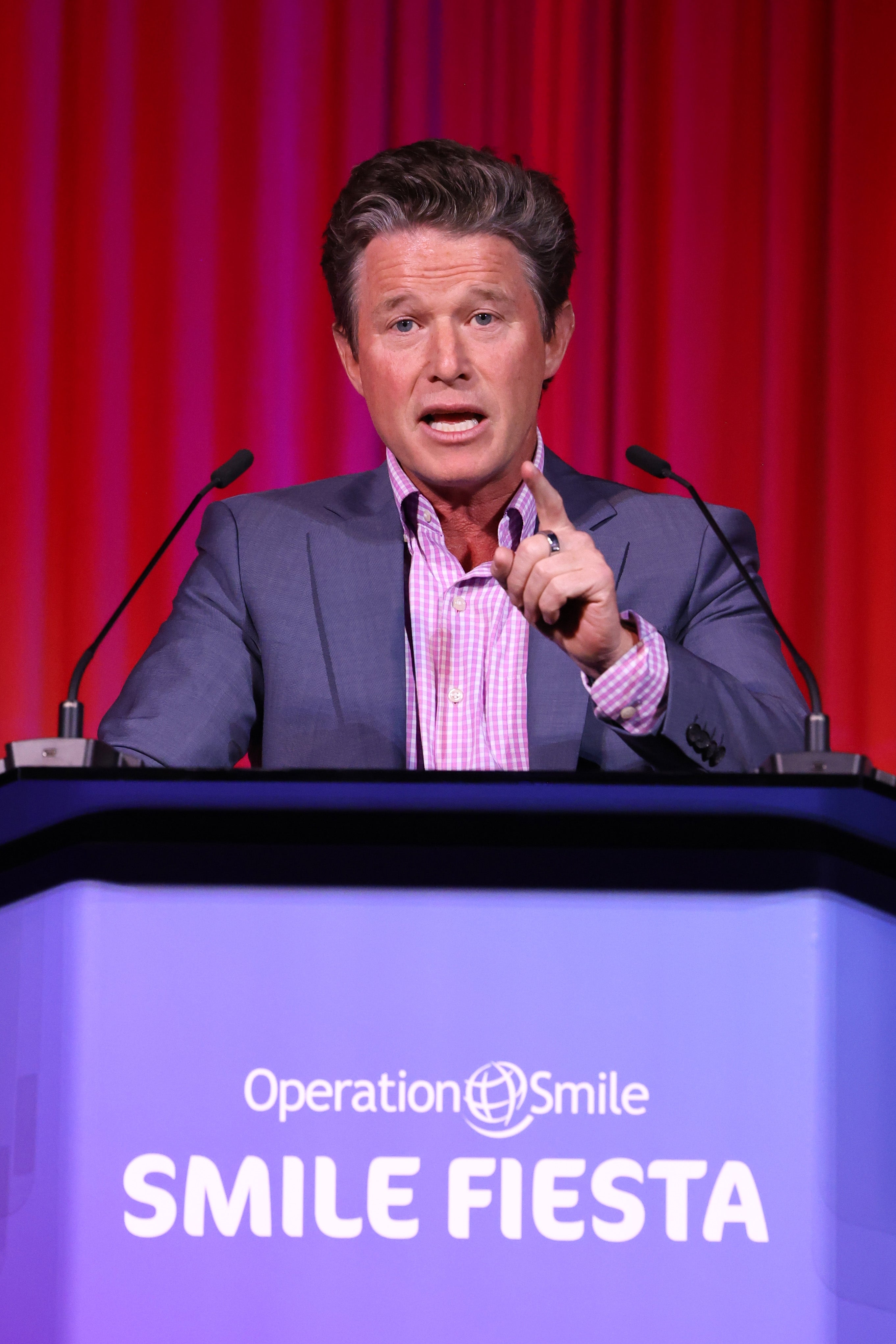 A idiosyncratic speaks passionately astatine a podium labeled "Operation Smile Smile Fiesta," wearing a suit and checkered garment against a reddish curtain backdrop