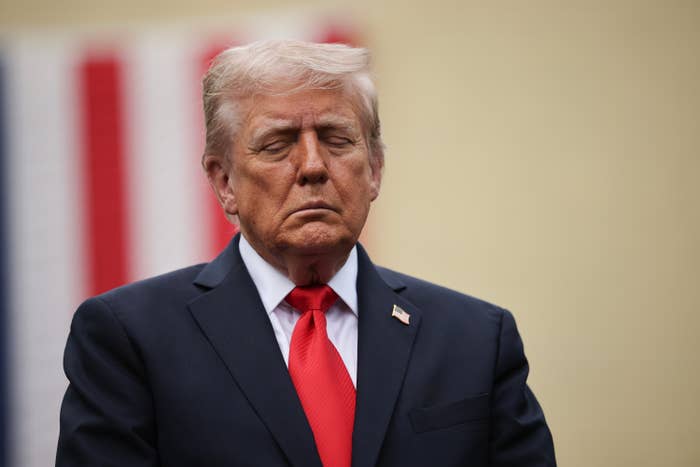 A idiosyncratic   successful  a suit   and reddish  necktie  stands with eyes closed, appearing contemplative, with an indistinct emblem  background