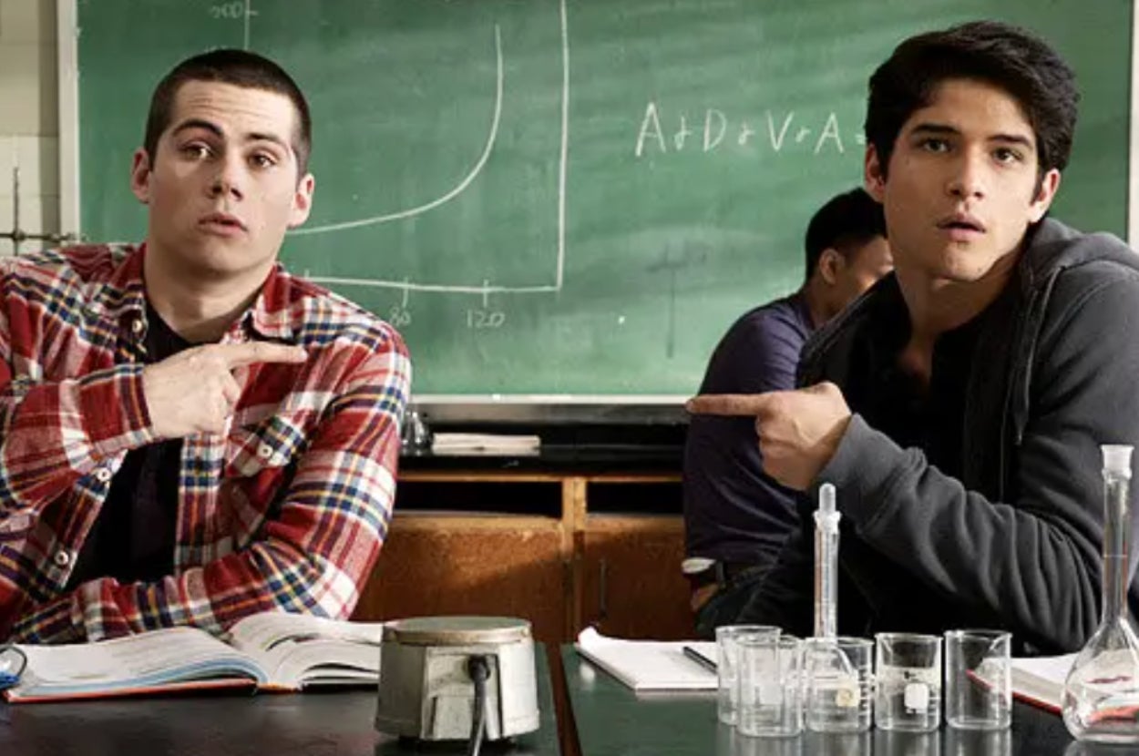 Two young men in a classroom are pointing in opposite directions, seated in front of desks with lab equipment and an equation on the chalkboard