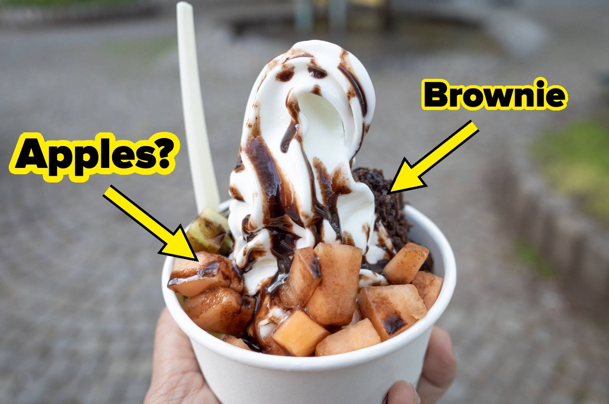 A hand holding a cup of soft serve ice cream topped with chocolate drizzle, pieces of fruit, and brownie, with arrows pointing to toppings