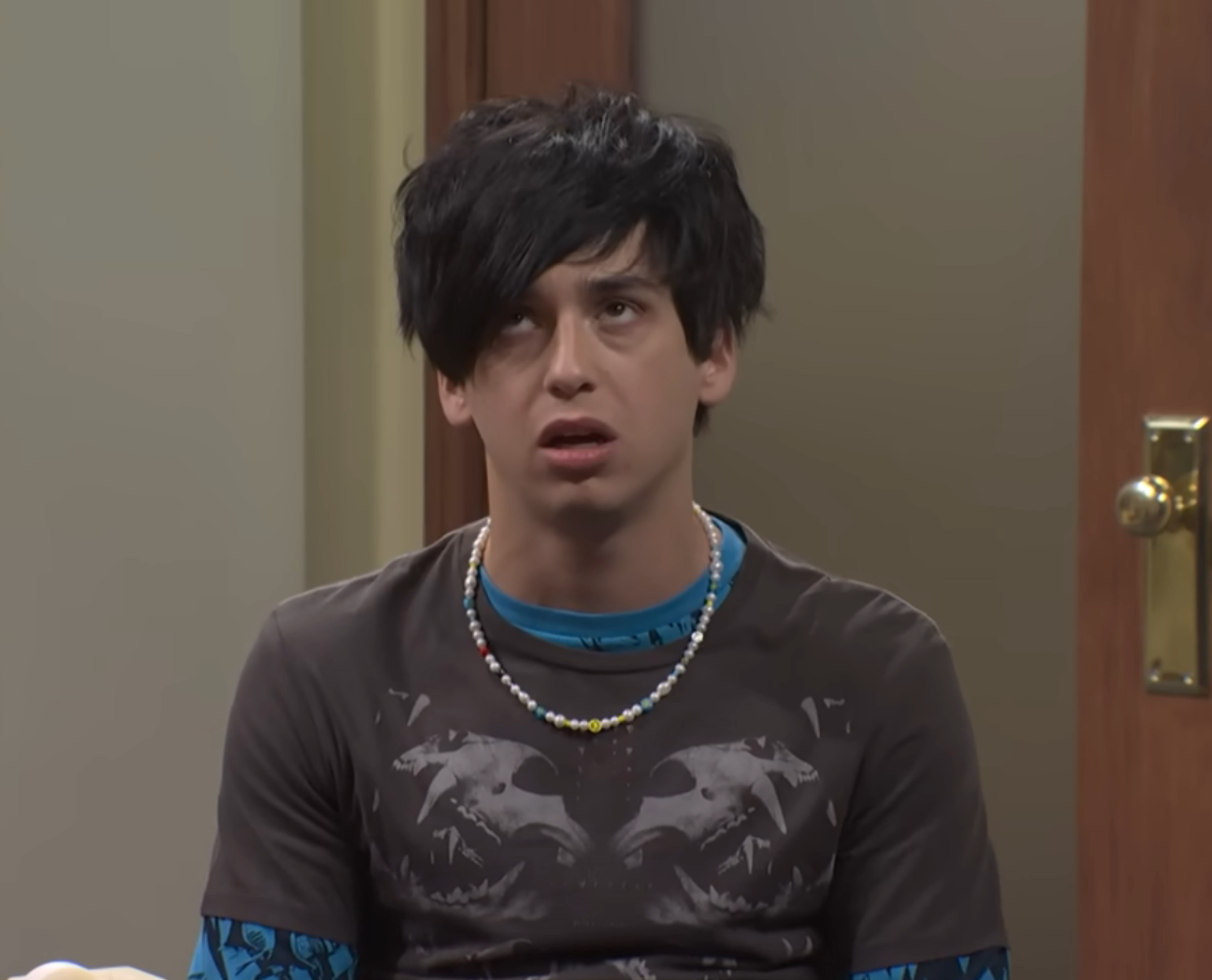 Person wearing a casual necklace and graphic t-shirt appears tired or bored, with disheveled hair, sitting indoors