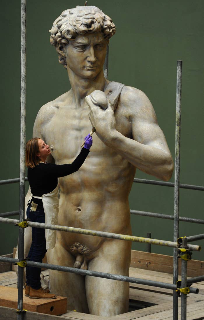 A woman in protective gloves carefully cleans a large, detailed statue of a muscular male figure, surrounded by scaffolding
