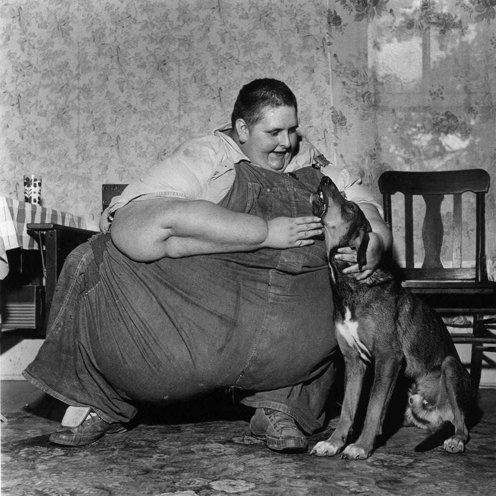A smiling person crouches to pet a dog in a warm room, surrounded by simple furnishings