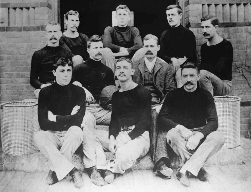 A historical black-and-white photo of a vintage sports team. Ten men in athletic attire or suits pose together on steps with basketball equipment nearby
