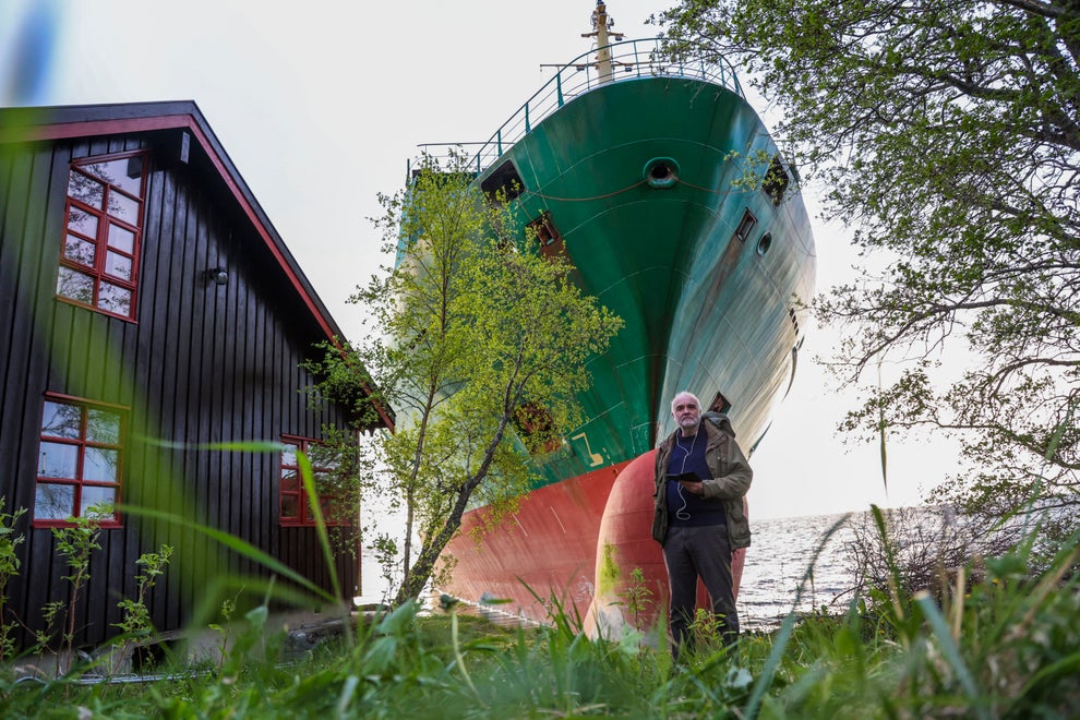A large ship sits on a grassy shore near a wooden building, with a person standing in front, holding a camera