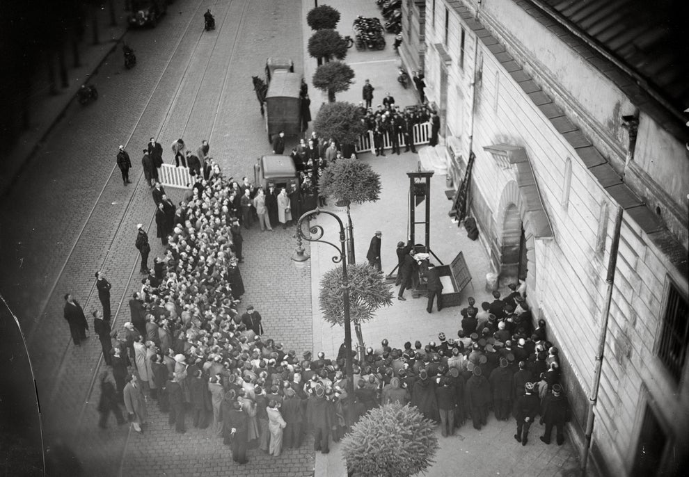 A historical black and white photo shows a large crowd gathered around a guillotine setup on a street, viewing an execution event