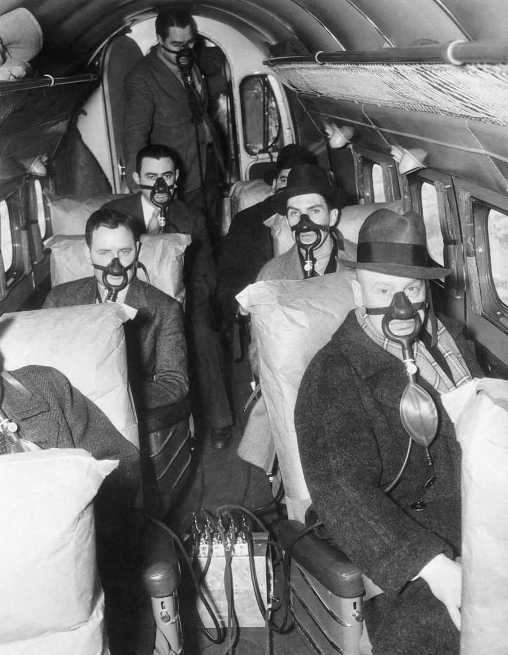 Passengers in an airplane cabin wearing oxygen masks during a safety demonstration in the early 20th century