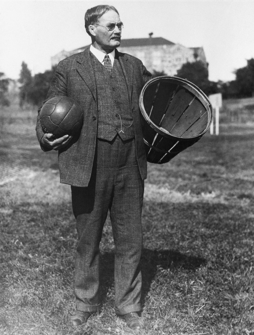 A historical figure in a suit holds an early basketball and a basket on a field, representing the origins of basketball