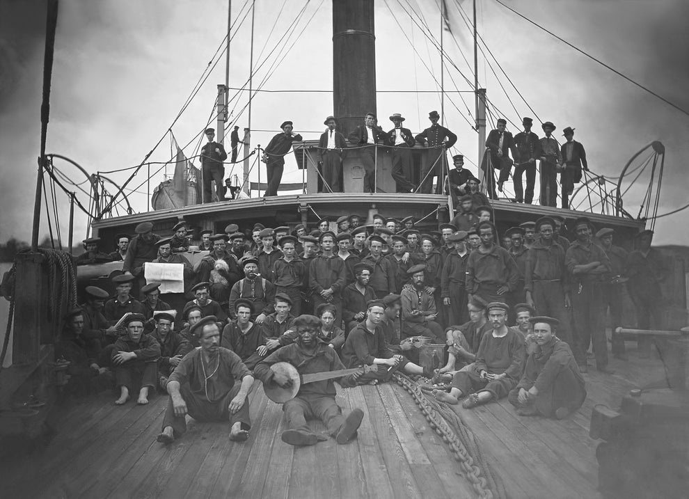 A large group of sailors dressed in uniforms posing on the deck of a ship with a sailor holding a guitar in the foreground