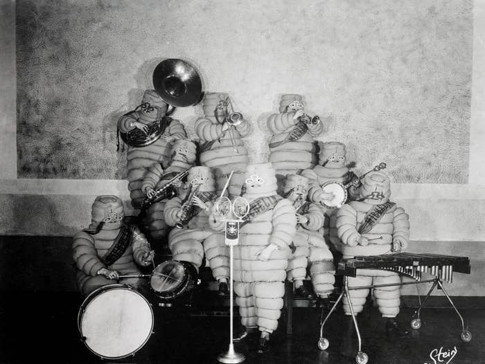 A group of people dressed as Michelin Men play various musical instruments on stage in a vintage black and white photo