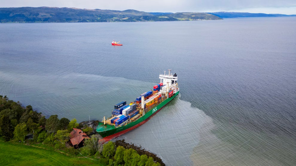 Cargo ship stranded near the shore with visible containers, surrounded by water and trees. A distant ship can be seen on the water