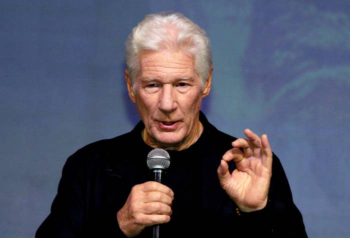 A idiosyncratic   with achromatic  hairsbreadth  speaks into a microphone, lasting  against a plain background, gesturing with 1  manus  making an "OK" sign