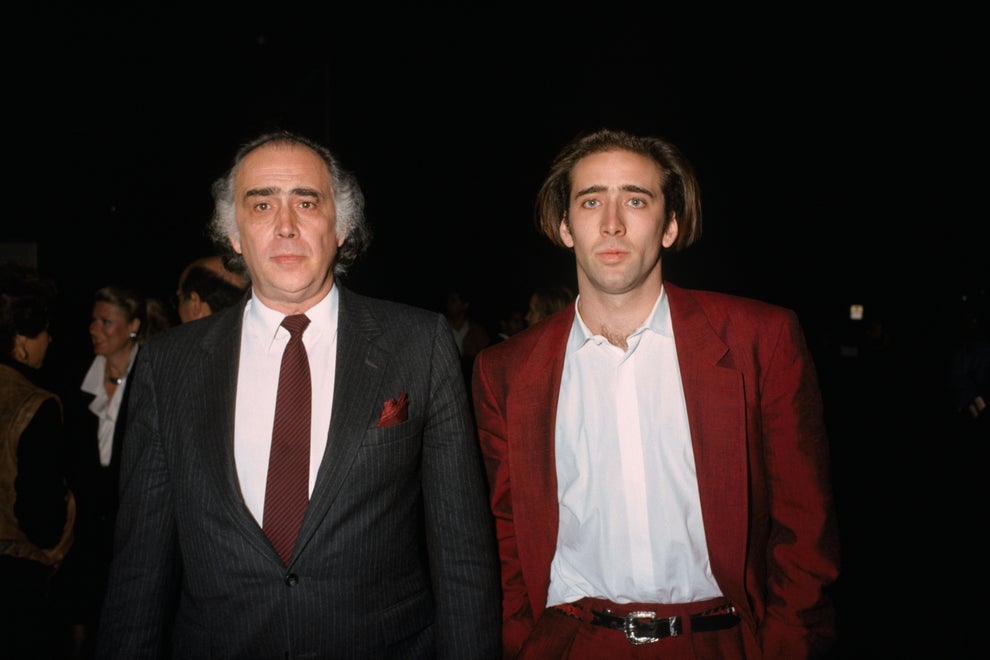 Two men at an event; the man on the left wears a suit with a tie, and the man on the right wears a casual open shirt and blazer