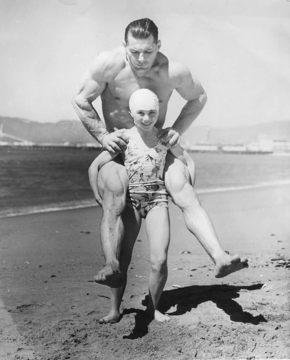 A muscular man gives a piggyback ride to a woman in a swim cap and swimsuit on a beach