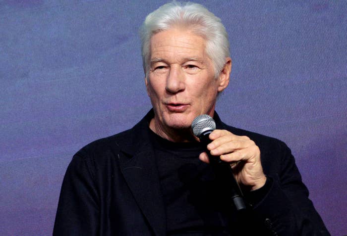 Older antheral   speaking into a microphone, wearing a acheronian  suit   overgarment   and shirt