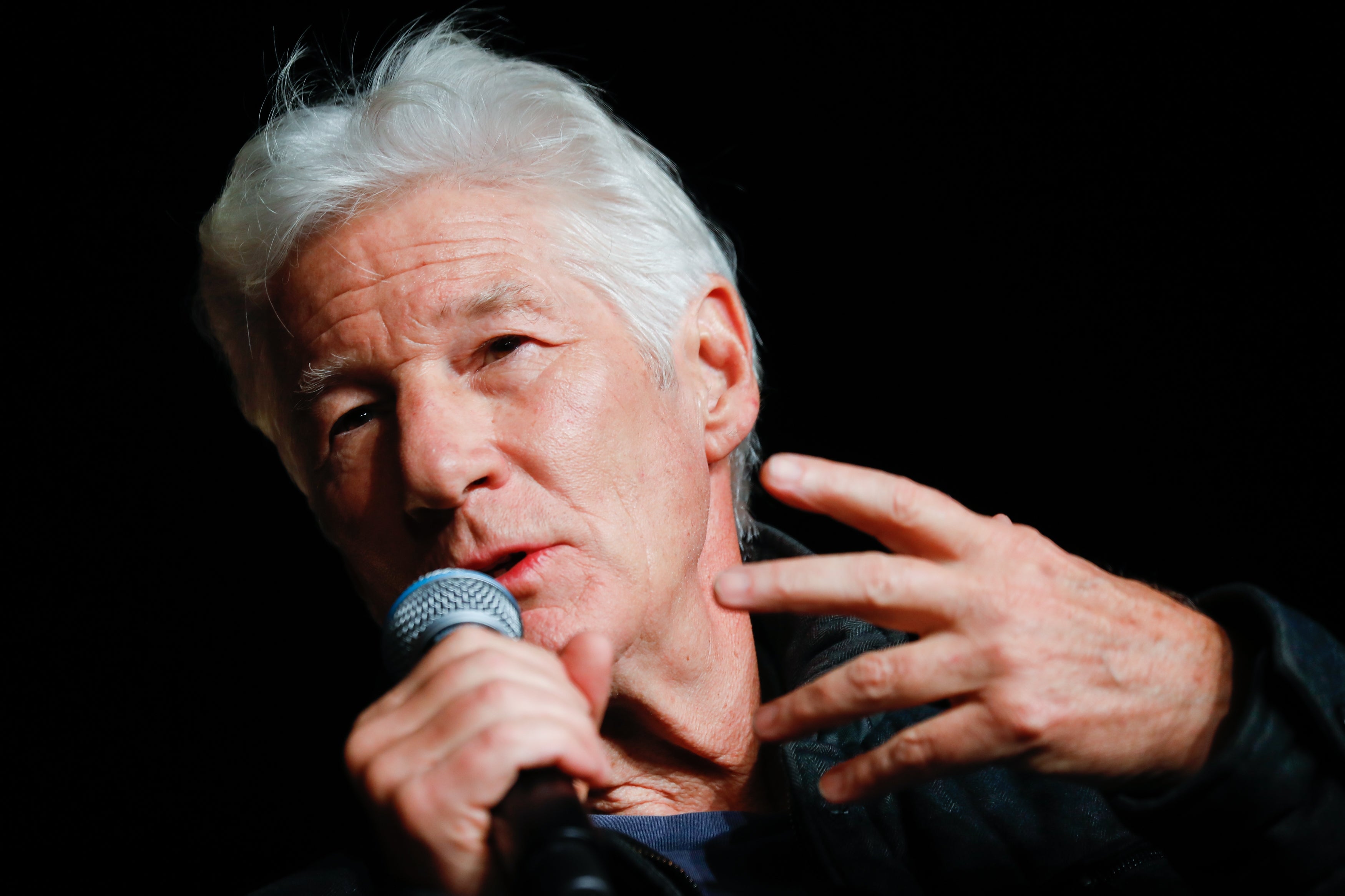 A idiosyncratic   speaks into a microphone, gesturing with 1  hand, wearing a acheronian  jacket