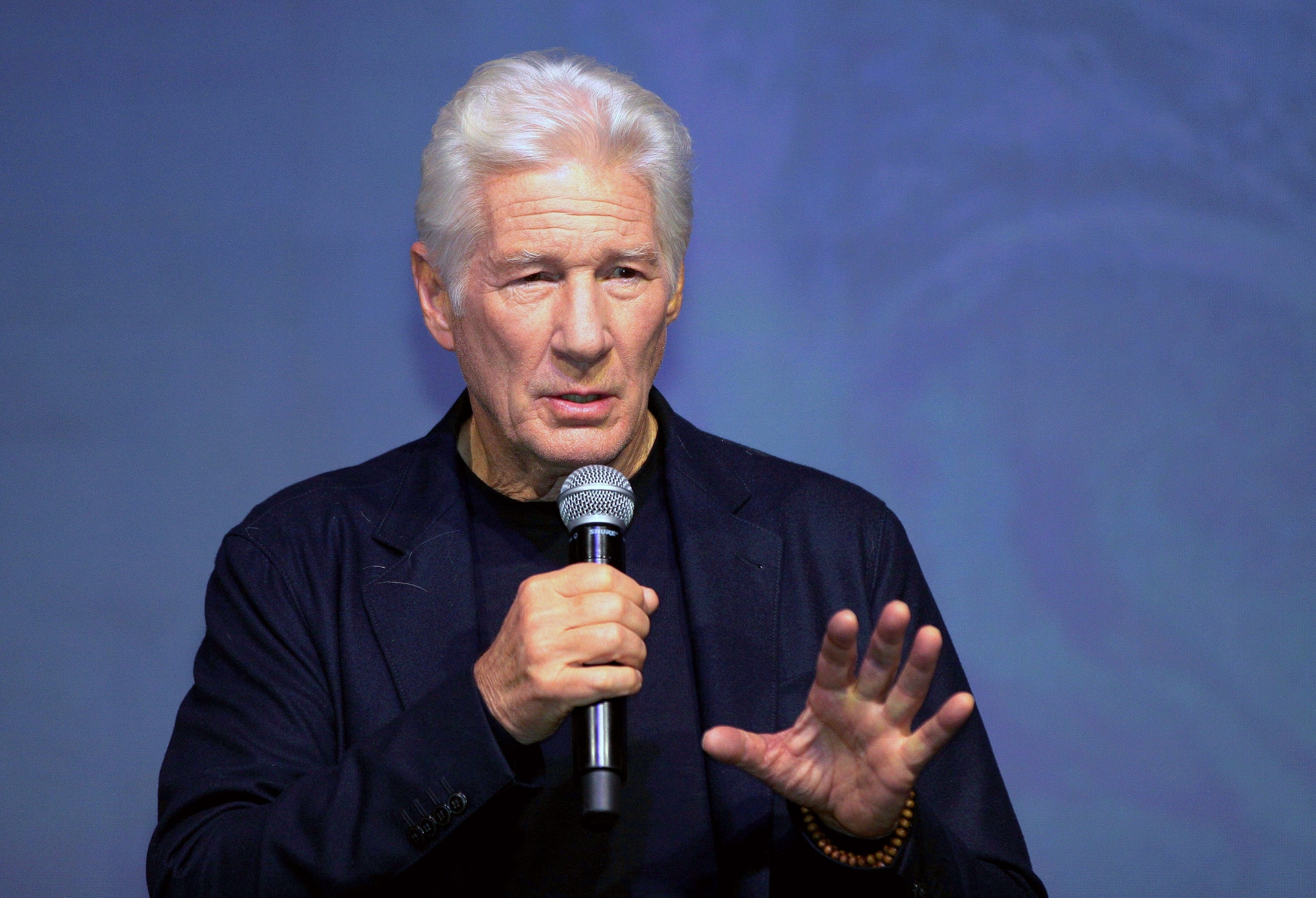An older antheral   speaks into a microphone connected  stage, wearing a acheronian  suit   with a bracelet connected  his near  wrist