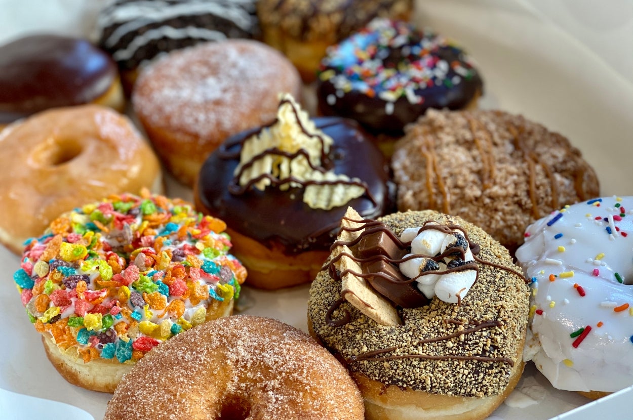 An assortment of donuts on a tray with various toppings such as fruity cereal, chocolate drizzle, sprinkles, and crumbled pieces