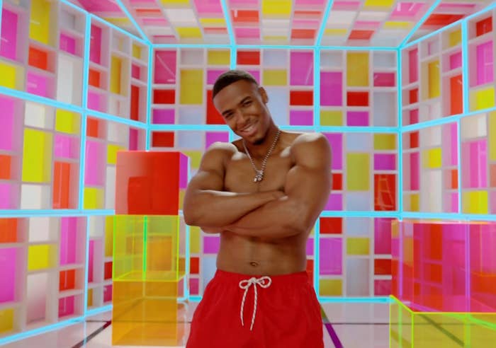Man with abbreviated  hair, shirtless successful  reddish  shorts, smiles with arms crossed successful  a colorful, geometric room