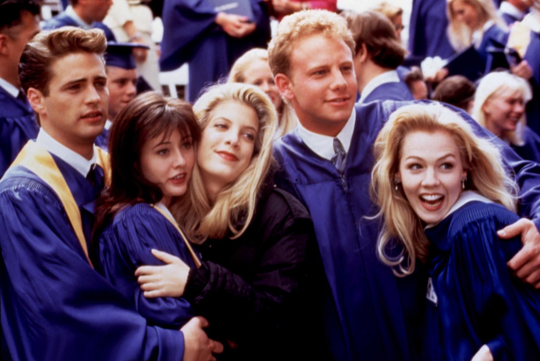 Actors in graduation robes pose closely together at a ceremony, smiling and celebrating