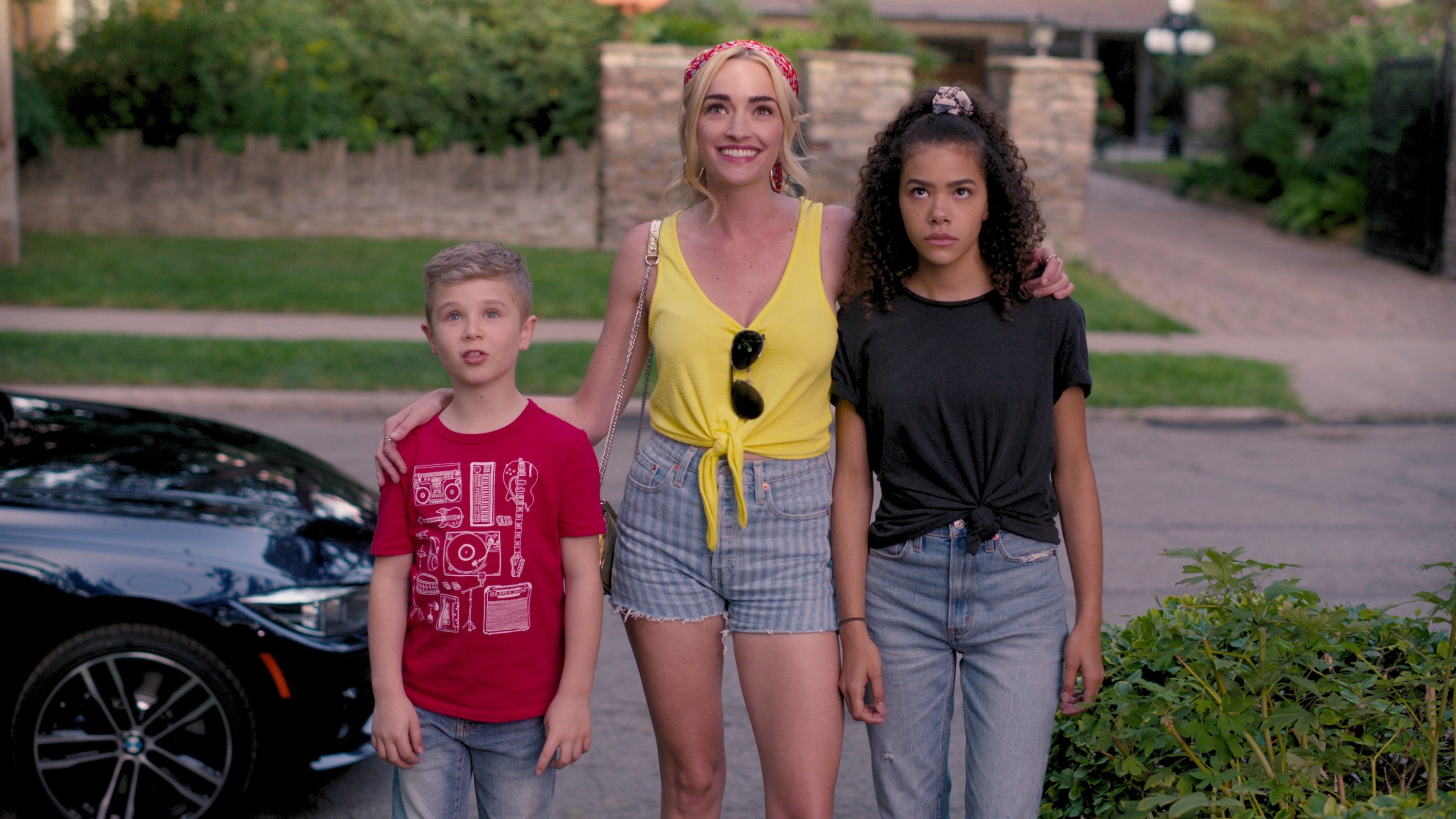 Three people stand in a suburban street: a smiling woman in shorts and a tank top with two children, one on each side, looking at the camera