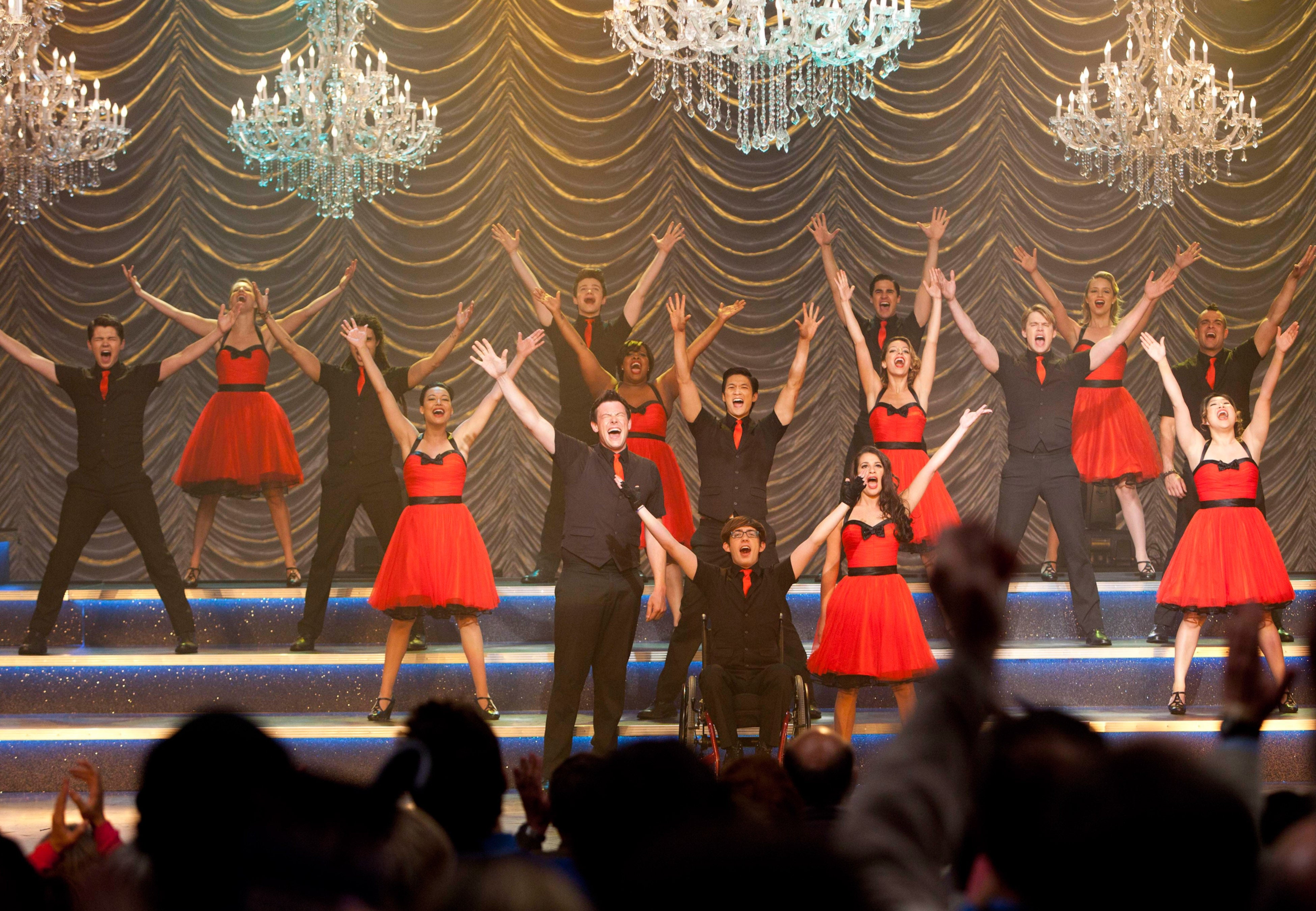 A vibrant stage performance with a group of singers and dancers in coordinated outfits, arms raised, under chandeliers. Audience members visible