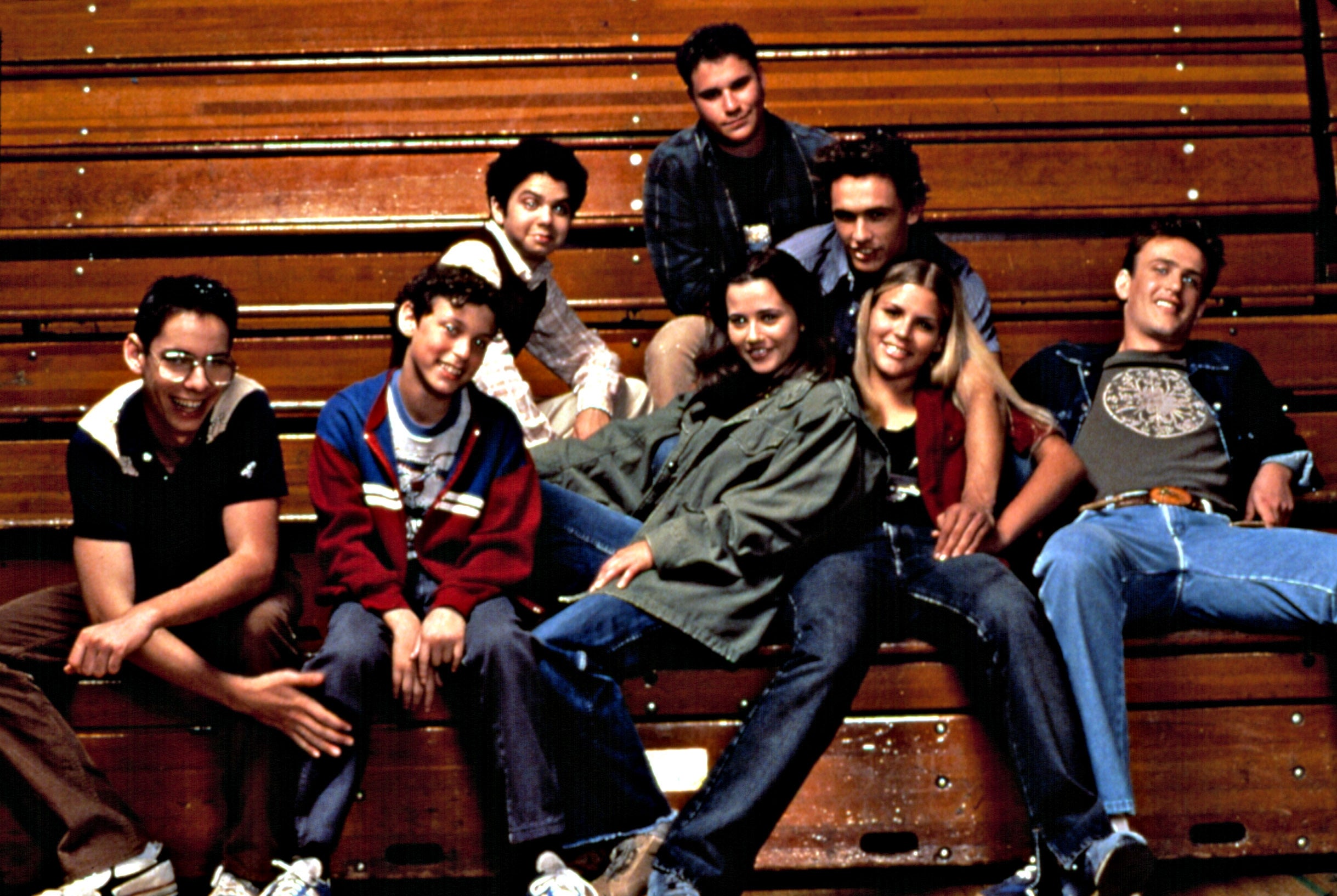 A group of young adults in casual '90s fashion sit on bleachers, smiling