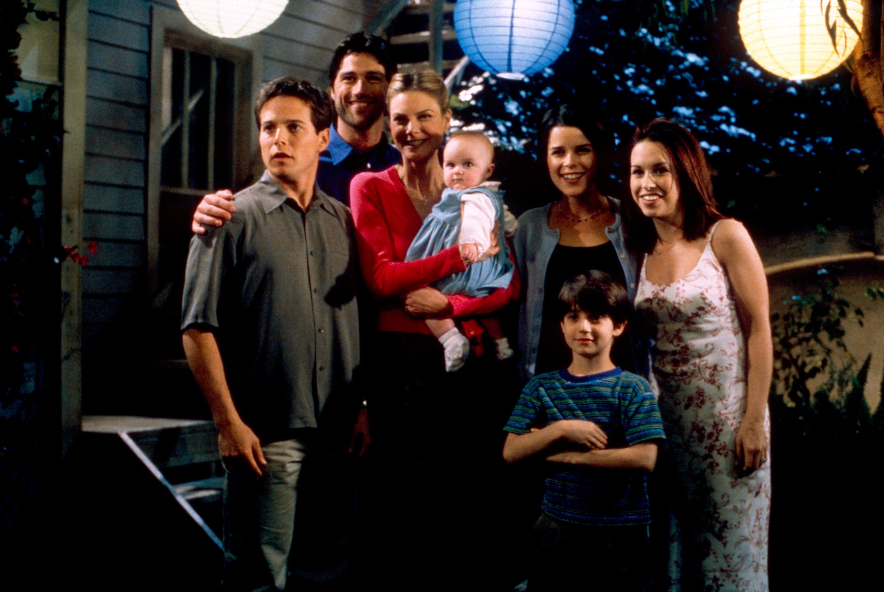 Group of actors posing outdoors under lanterns, featuring a woman holding a baby. Everyone is casually dressed, smiling, and standing together