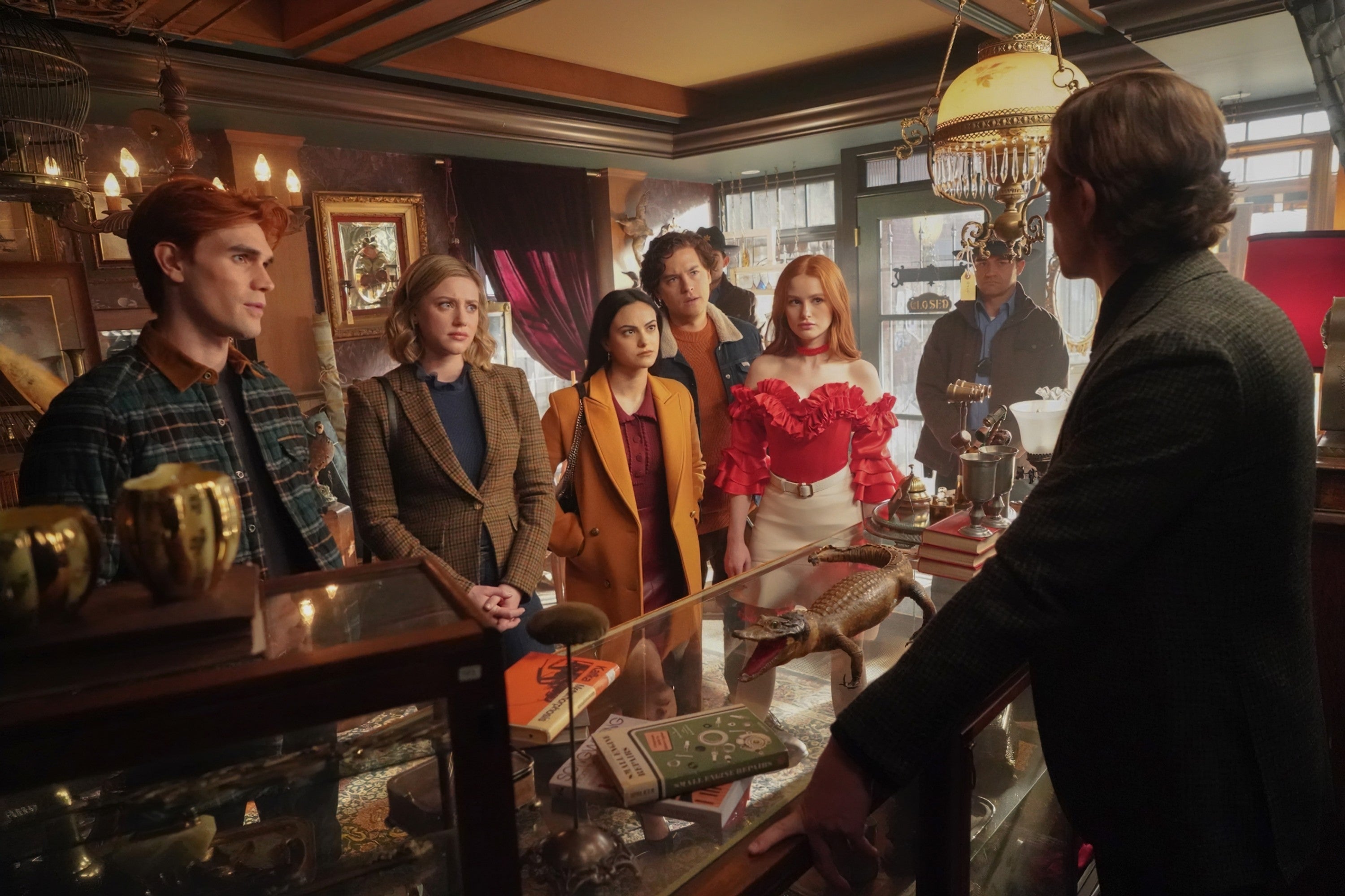 Cast of a TV show stands in a vintage shop, engaged in conversation with a shopkeeper. One character wears a red ruffled top with white pants