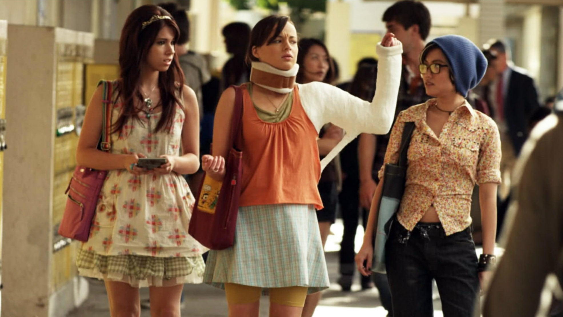 Three young women walk in a school hallway. One wears a neck brace and cast, pointing forward. They appear to be chatting animatedly