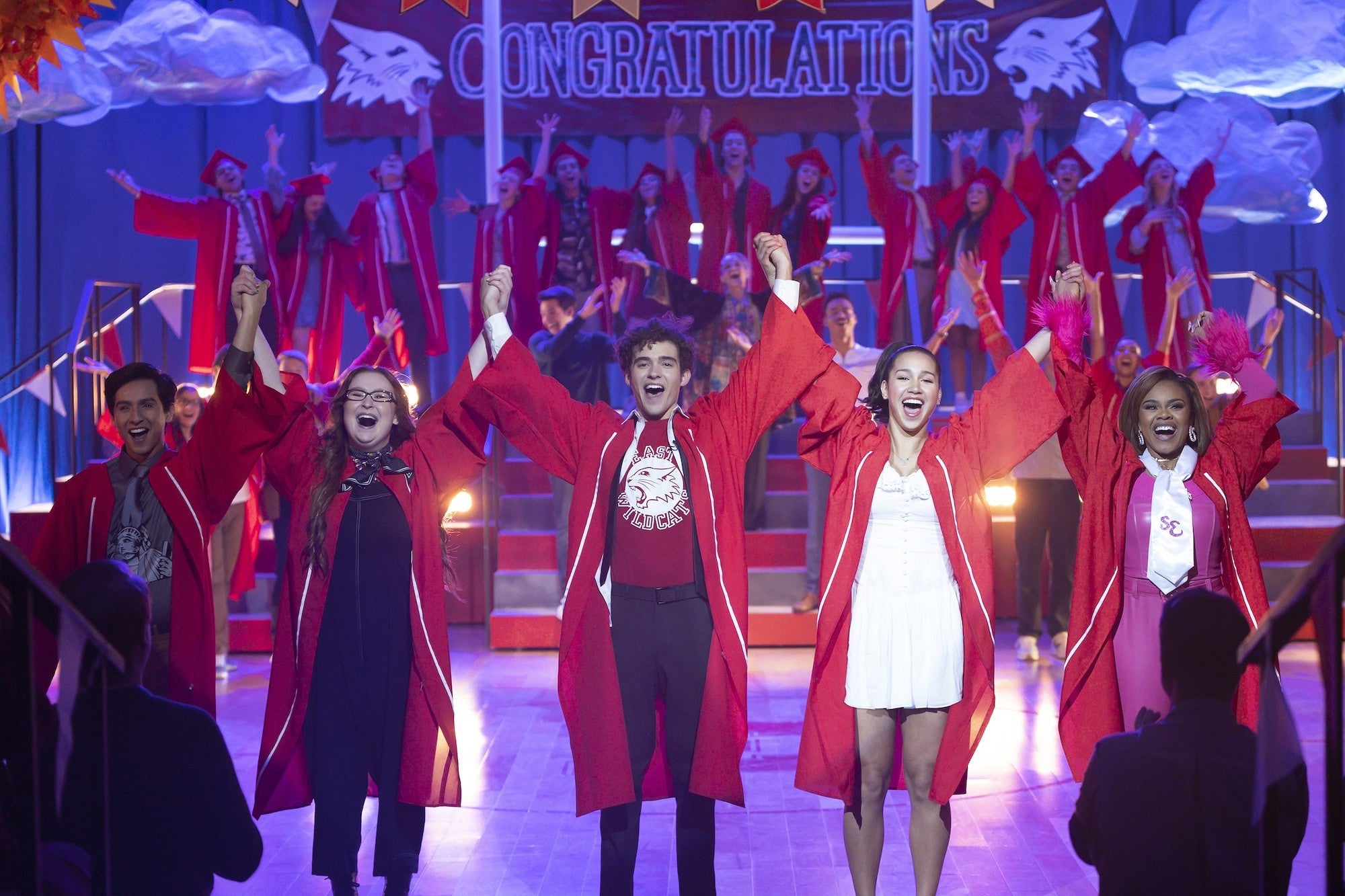 People in red graduation robes celebrate on stage with raised arms, under a "Congratulations" banner, suggesting a joyful performance or ceremony