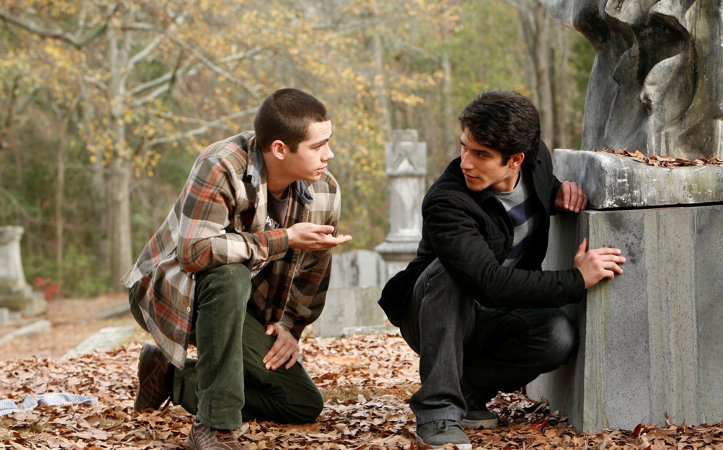 Two actors in a cemetery scene, one crouching in a plaid shirt, the other in a jacket, both appearing concerned. Fallen leaves cover the ground