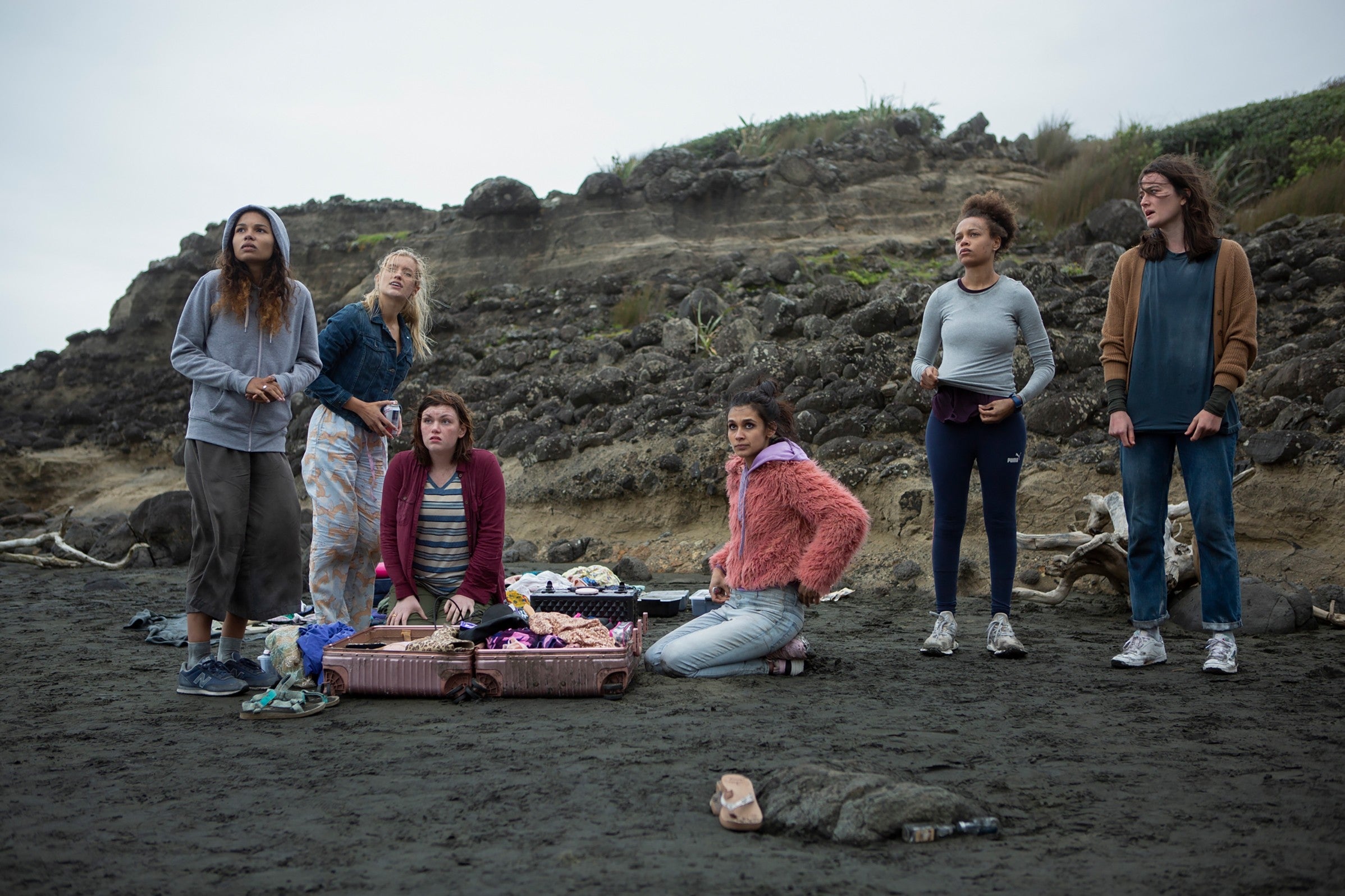 Six people on a beach, looking worried. Suitcase open with personal items. Rugged terrain in background, giving a sense of isolation