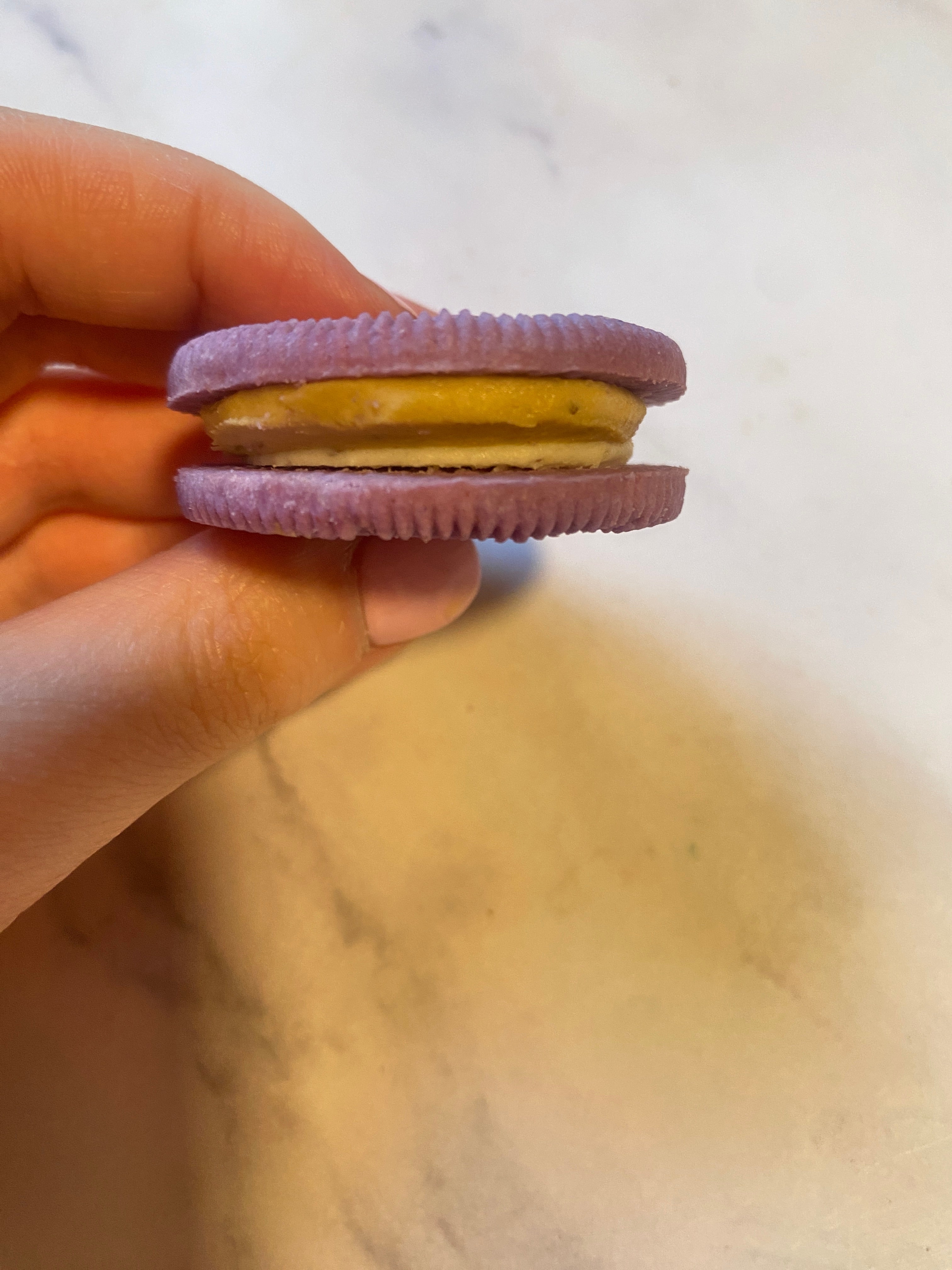 A hand holds a sandwich cookie with purple outer layers and a yellow cream center against a neutral background