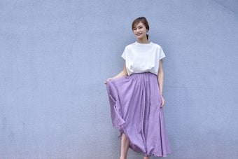 女性がシンプルな白のトップスとロングスカートを着て笑顔で立っている。
