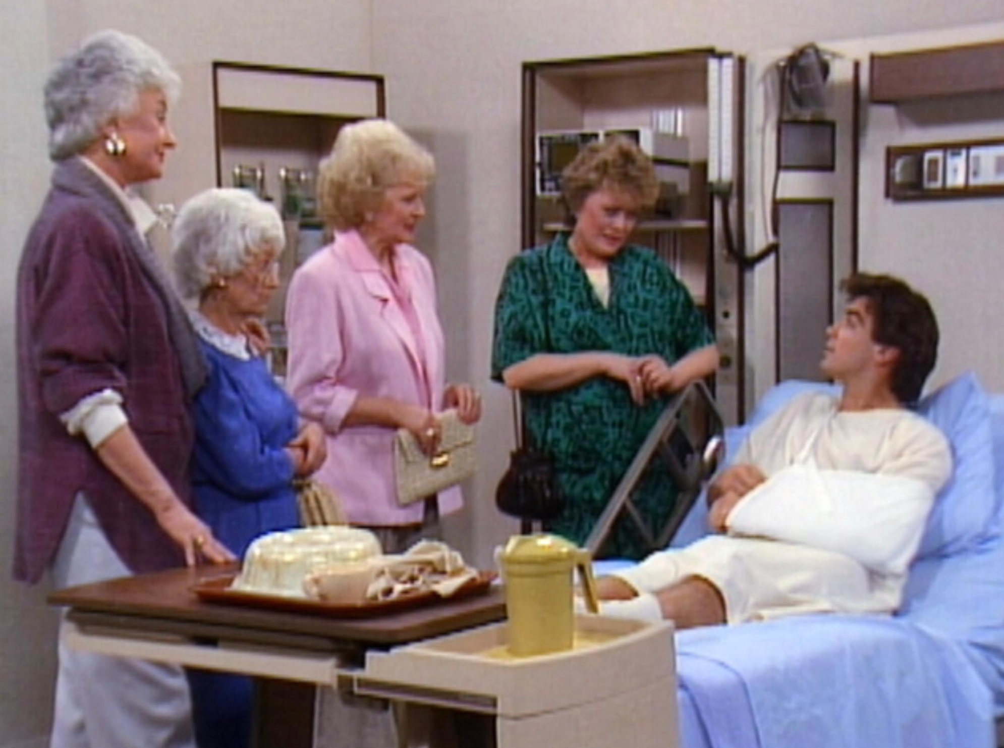 A diligent with an limb formed successful a infirmary furniture talks with 4 older women, each dressed successful casual 1980s-style clothing, gathered astir the bed