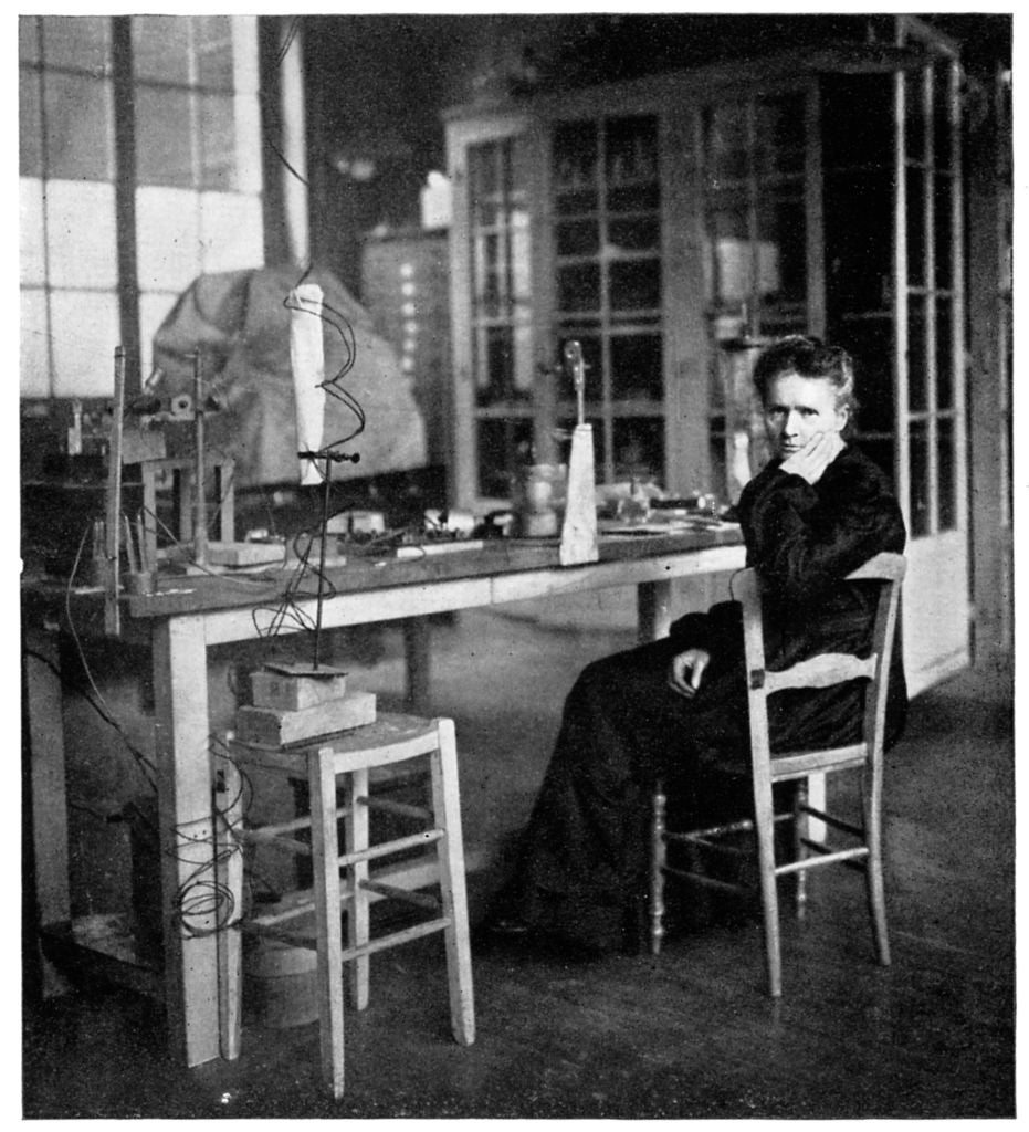 Person seated in a lab with scientific equipment on a wooden table, surrounded by shelves and cabinets