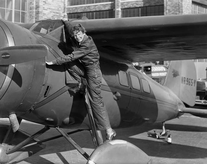 A person in a flight suit climbs onto the wing of a vintage aircraft, suggesting a historical aviation context