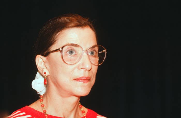 A person wearing large round glasses, red earrings, and a red top, with hair tied back, appearing thoughtful against a dark background