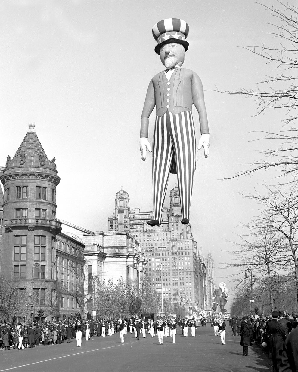 Giant Uncle Sam balloon floats above a parade with onlookers and vintage buildings in the background