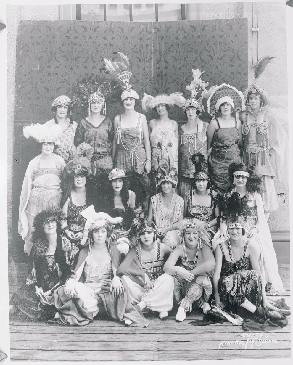 Vintage group photo of 19 women in elaborate, feathered costumes from early 20th century, posed together on a wooden floor