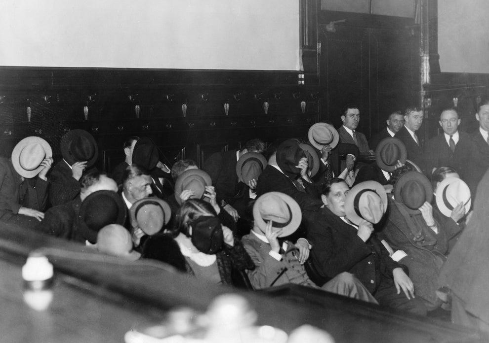 Historical black-and-white photo of people sitting indoors, hiding their faces with hats