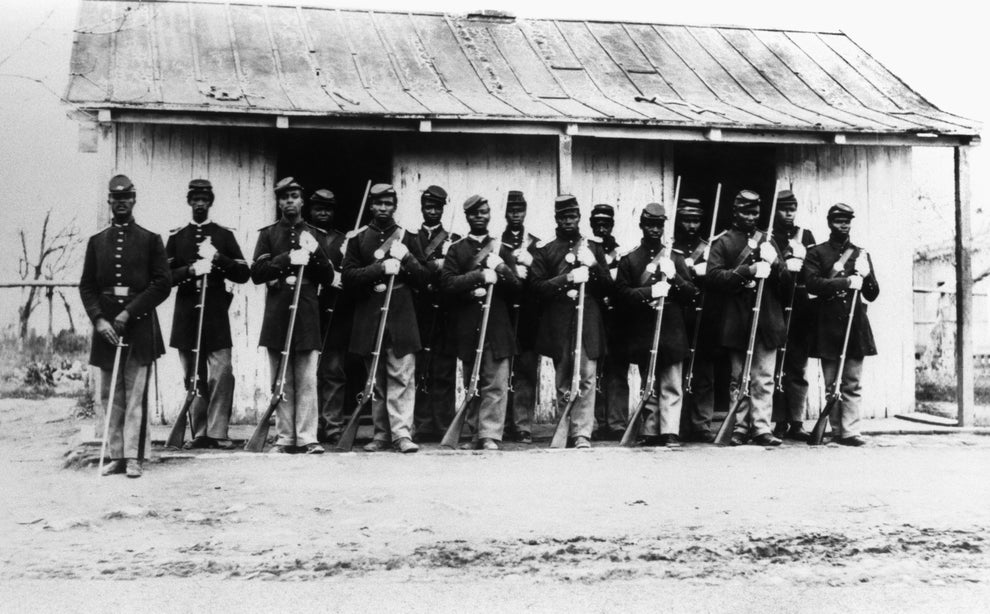 Historical photograph of a group of African American soldiers in uniform with rifles, standing in front of a wooden building