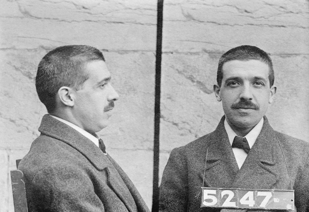 Historic mugshot of a man with short hair and a mustache, wearing a suit and overcoat, facing forward and in profile