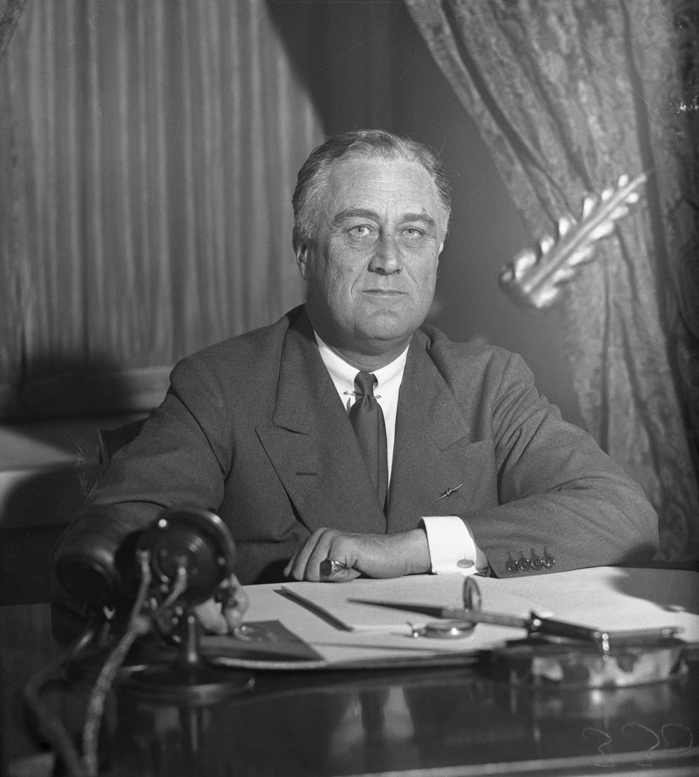 A historical figure in a formal suit sits at a desk with papers and a telephone, suggesting a setting of authority and leadership