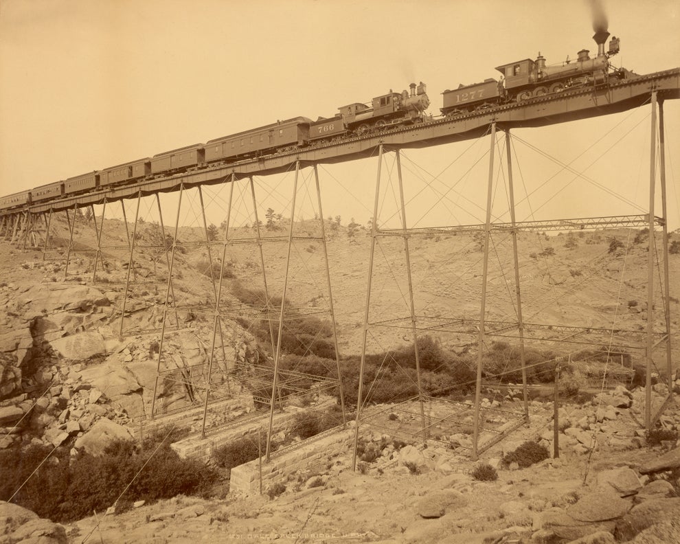 Historic train crossing a tall, narrow trestle bridge amidst a rocky landscape, steam billowing from the locomotive