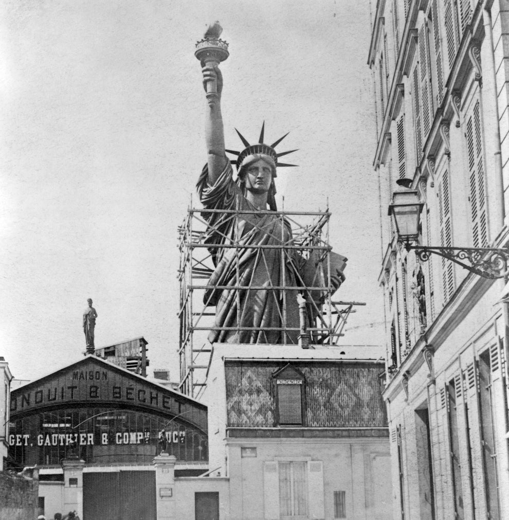 Statue of Liberty under construction in France, with scaffolding around the statue, situated between buildings