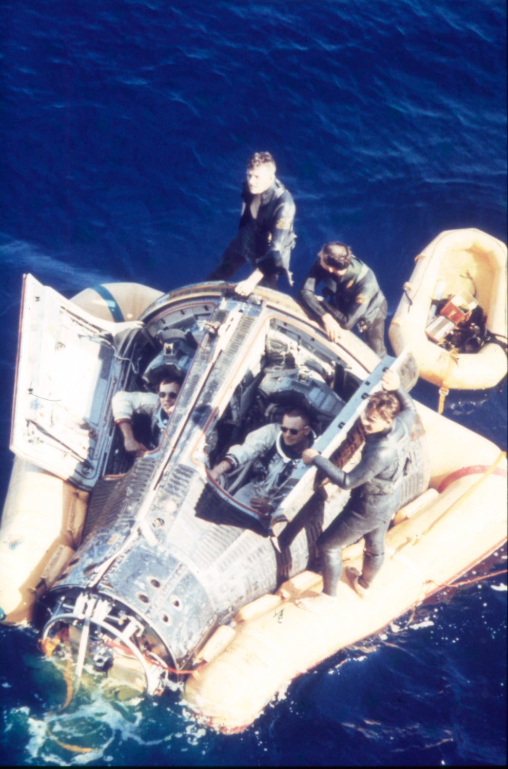 Astronauts in a life raft with a space capsule floating on the ocean after a splashdown, surrounded by crew members