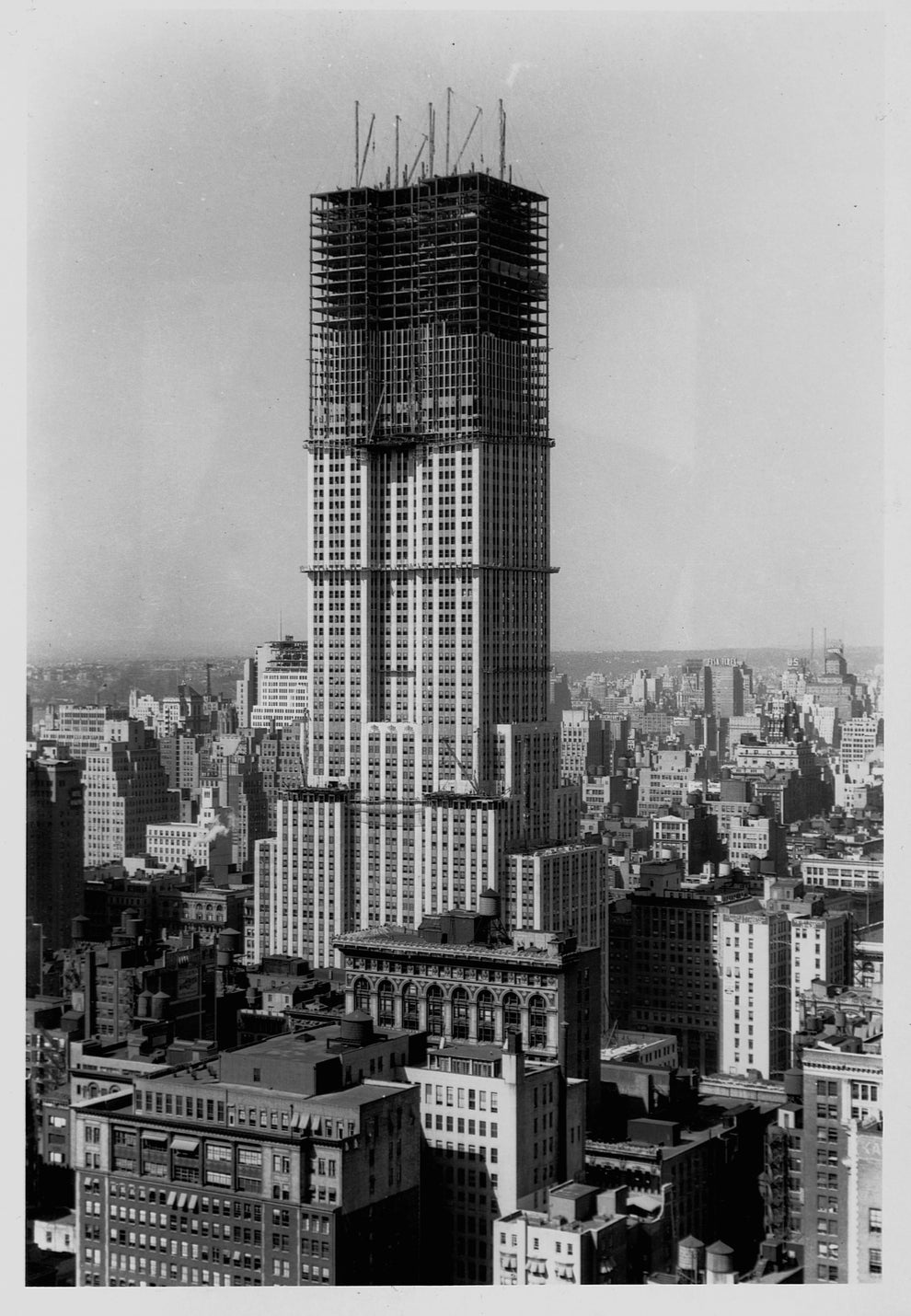 The Empire State Building under construction, surrounded by other buildings, scaffolding visible at the top. Urban skyline in the background